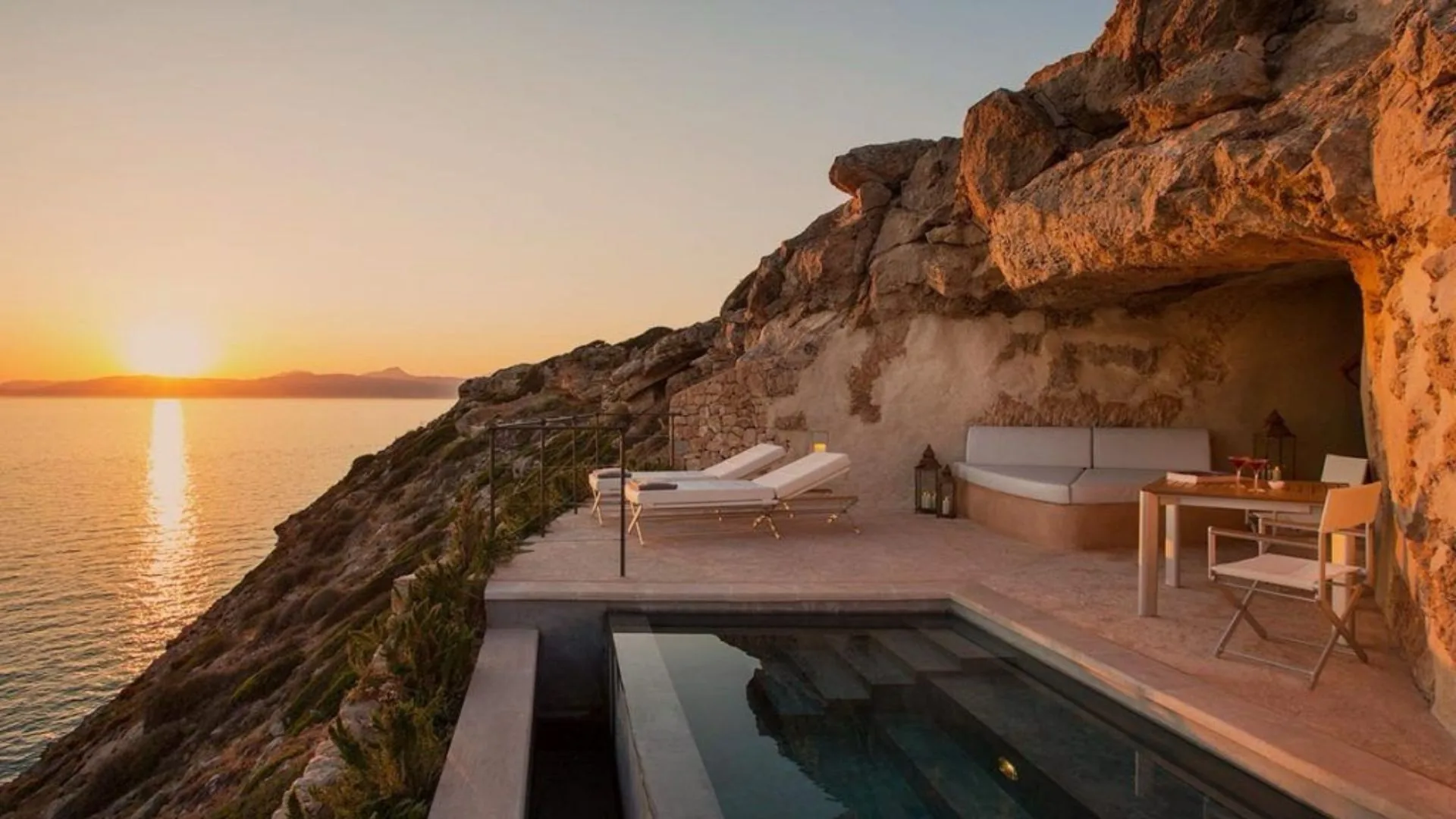 Seaside terrace with a private pool beneath a rocky overhang at sunset.